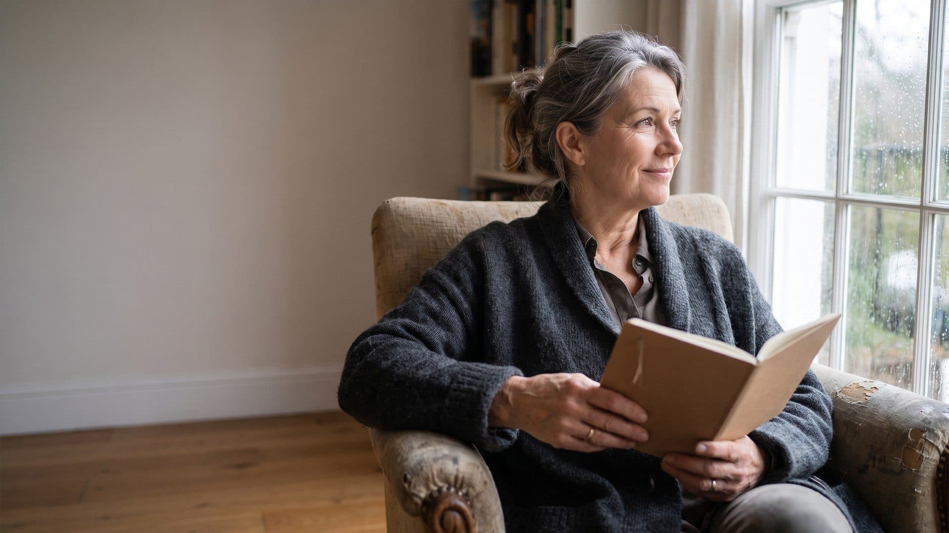 A woman with grey hair sitting in an armchair by a rainy window, holding an open book and looking outside
